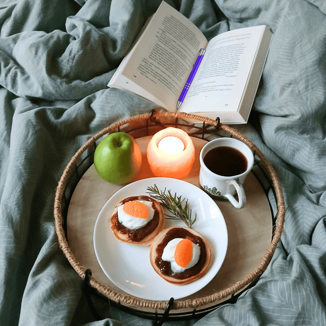 wellness flat-lay scene with candle, coffee, book, purple pen and healthy food on a plate, all sitting on a duvet