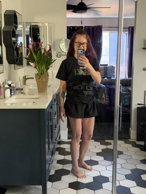 Me in a baggy t-shirt and shorts wearing my back brace, standing in front of a mirror.